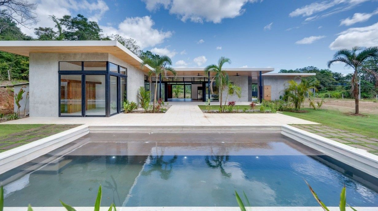 3D rendering of the interior of the social area of House CM, featuring a contemporary design featuring a wooden ceiling, floor-to-ceiling windows, and an open layout integrating the living, dining, and kitchen areas. The warm, natural finishes provide comfort, elegance, and a visual connection to the outdoor garden.