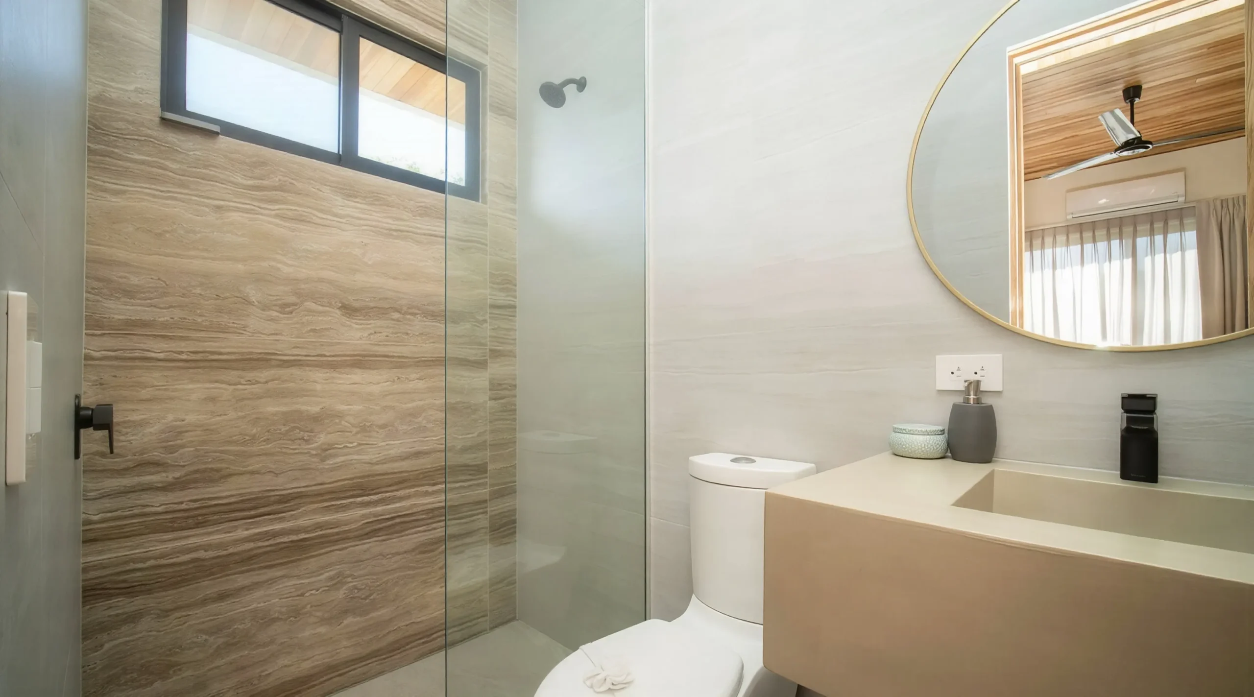 House Corona's sophisticated bathroom features a glass shower and natural stone walls, a modern sink, and an oval mirror. The window and ceiling fan bring light and freshness to this space.