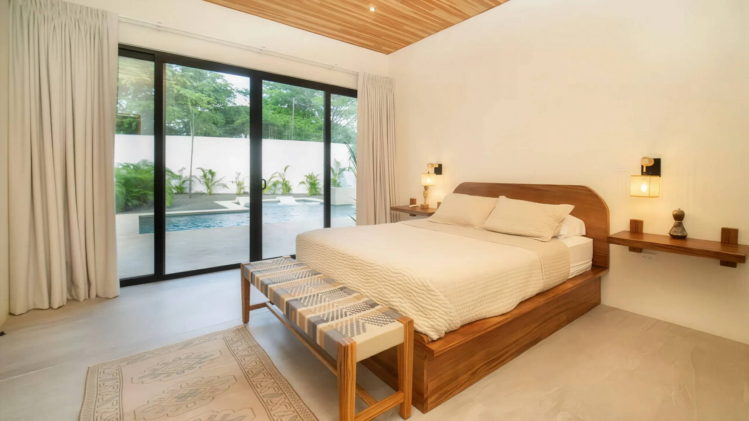 A cozy bedroom at House Corona features a wooden bed, wooden nightstand-style shelves, and a panoramic view of the pool through large windows.