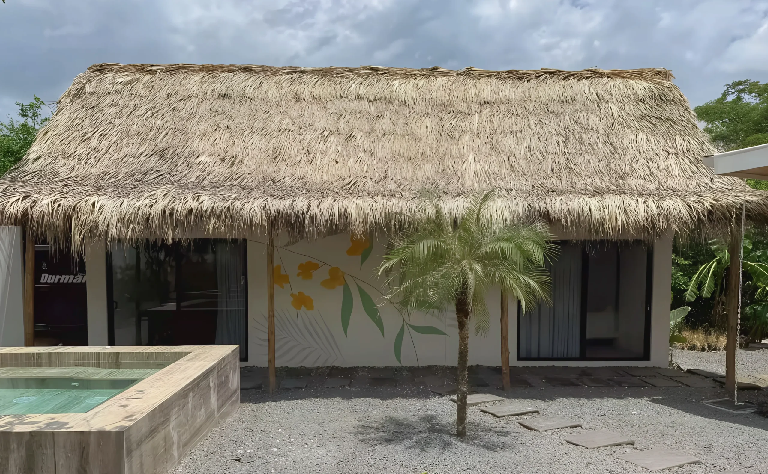 Exterior view of Casa Corona's Casita, a thatched cottage with palm trees and a small pool.