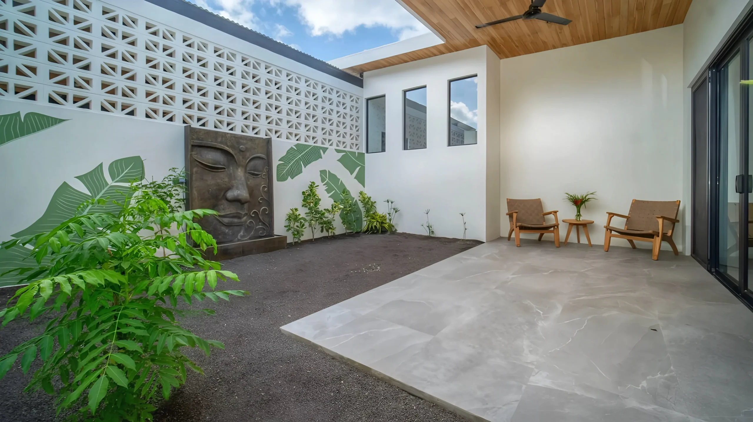 House Corona's inner terrace features a fountain with a carved face that blends with the lush vegetation. The space includes wooden chairs and a ceiling fan.