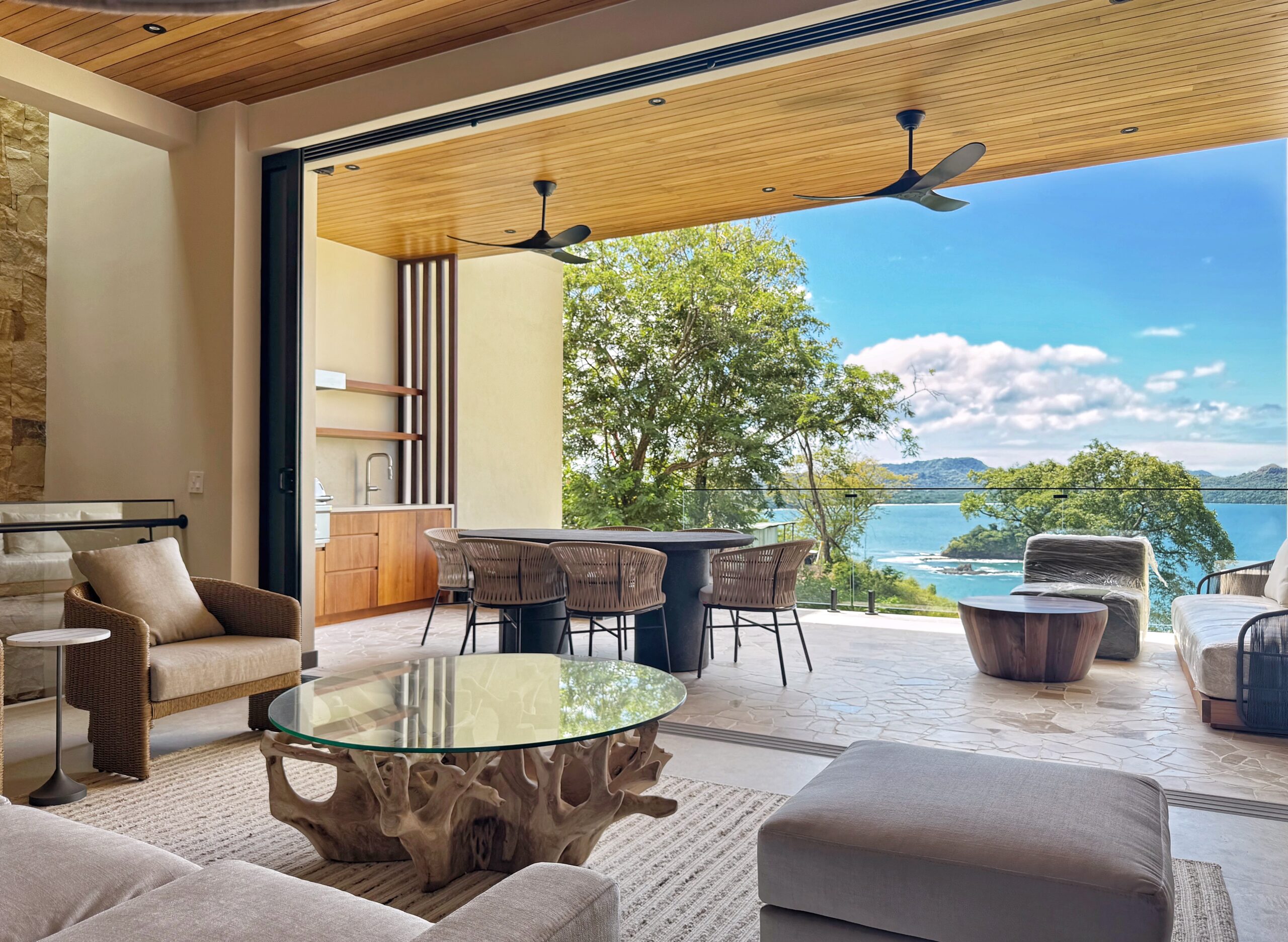 3D rendering of the open-plan living room of Model House overlooking the sea, featuring a comfortable white sofa, a wooden coffee table with plants, and a dining area with wicker chairs. The space features a wooden ceiling with ceiling fans, glass railings, and lush vegetation surrounding it.